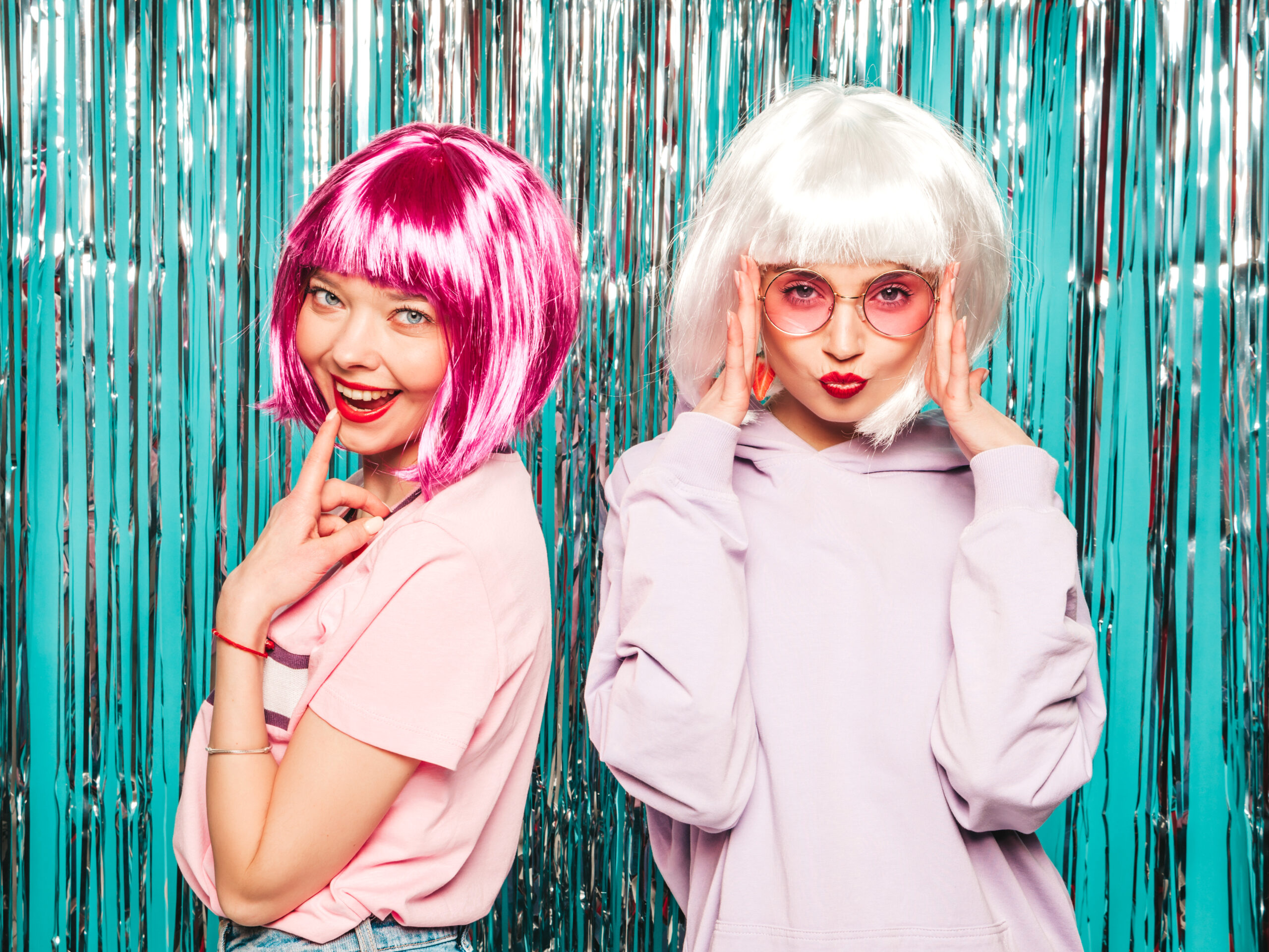 Two young sexy hipster girls in  wigs and red lips.Beautiful trendy women in summer clothes.Carefree models posing on blue silver shiny tinsel background in studio.Positive female having fun