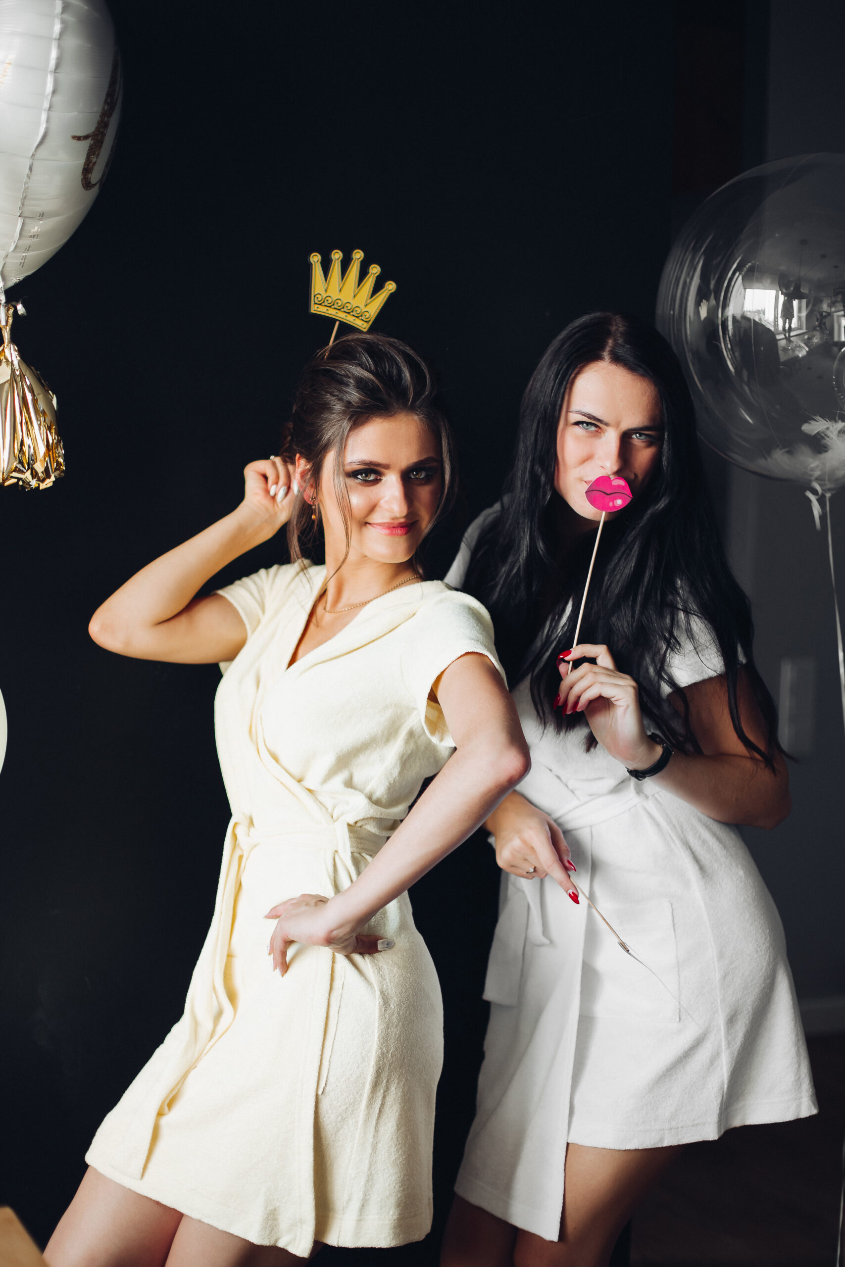 Two beautiful young ladies holding accessories and posing at camera. Girls in white and yellow dresses coming together to party chilling out. Queen of party with sad friend standing among decorations.